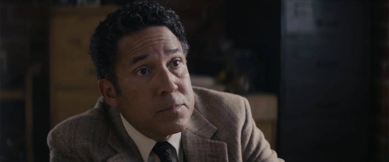 A Christmas Mystery (2022) movie still: closeup — A dimly lit office interior contains wooden furniture and a dark metal filing cabinet. An older…