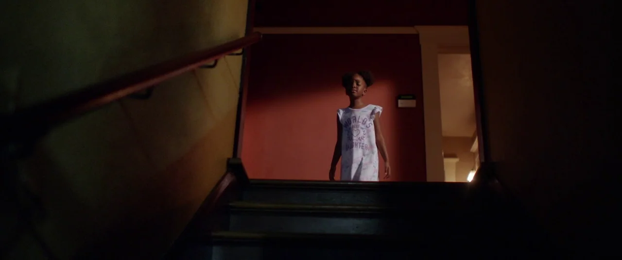 Blindfire (2020) movie still: medium — A dark staircase leads up to a dim corridor with deep red walls. A young girl stands on the stairs looking…