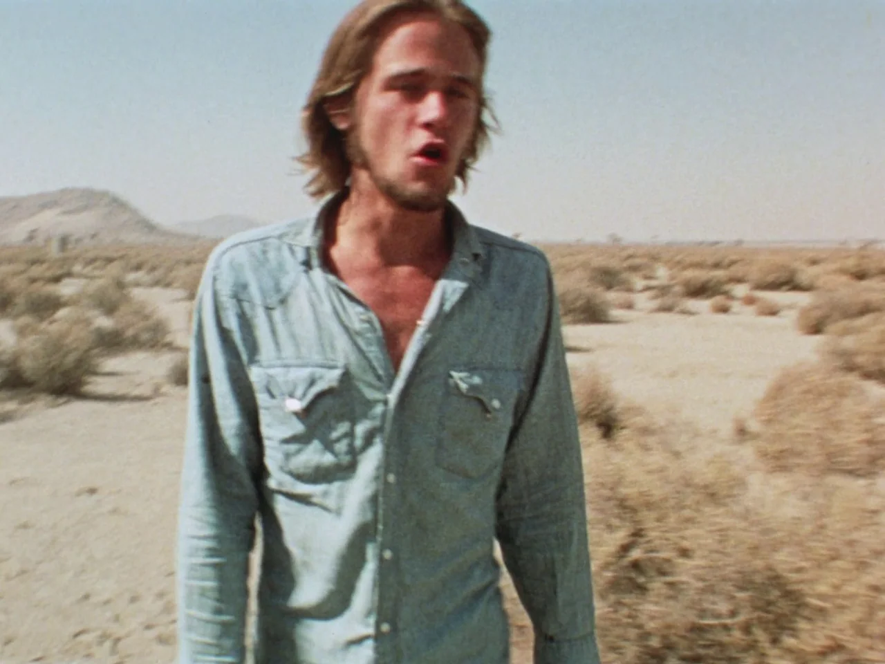 Punishment Park (1971) movie still: medium — A vast, dry desert landscape stretches into the hazy, arid distance. A young man with long hair walks…
