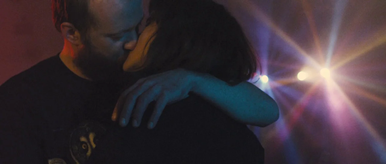 Sightseers (2012) movie still: medium — A dimly lit space features a couple kissing during a live music performance. Bright stage lights create…