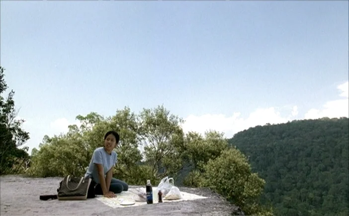 Blissfully Yours (2002) movie still: wide — A woman sits on a patterned blanket on a rocky cliff edge with greenery. She gazes out toward a large…