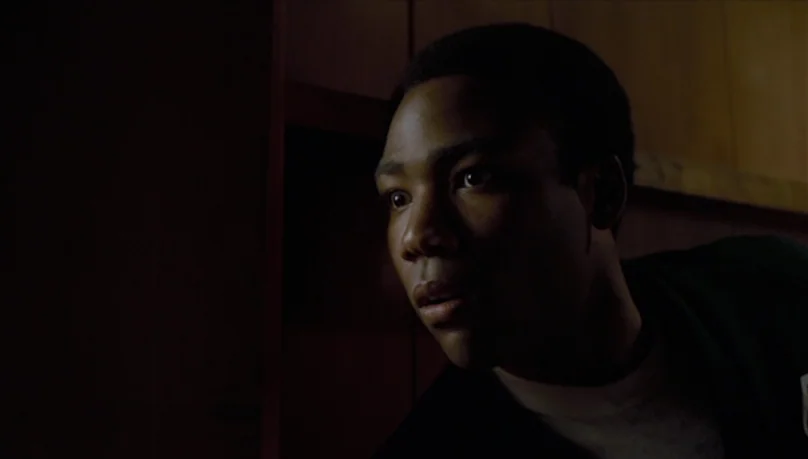 Mystery Team (2009) movie still: closeup — A dimly lit interior space with dark walls and minimal environmental detail. A young man stares intently…