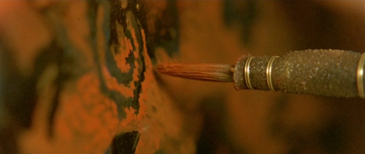 Girl with a Pearl Earring (2003) movie still: closeup — An artist's brush touches a textured surface covered in dark and reddish-orange paint. The…