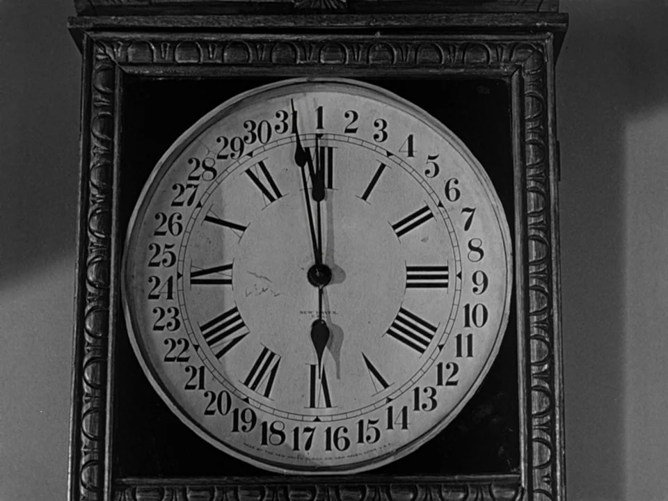 It’s A Wonderful Life (1946) movie still: closeup — A vintage wooden clock hangs on a wall with Roman numerals and numerical dates. The clock hands…