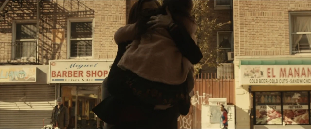 Exposed (2016) movie still: medium — An urban street with two storefronts featuring brick facades and signage for businesses. A person stands in the…