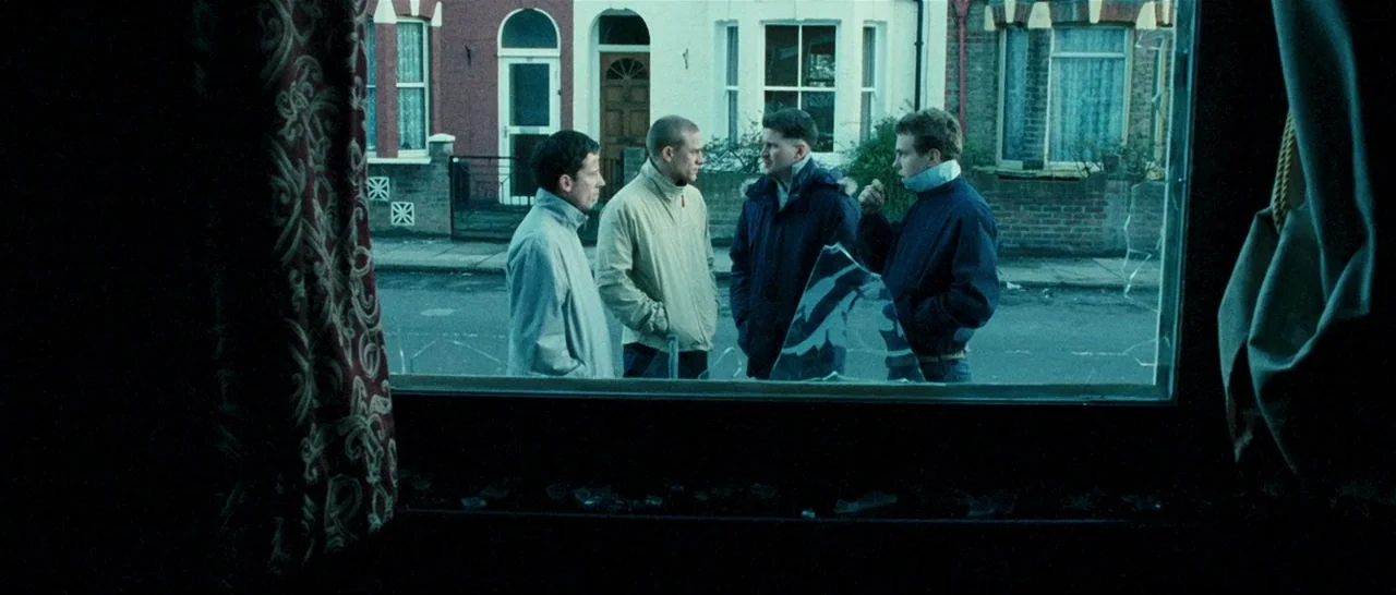 Green Street (2005) movie still: medium — Four men stand on a sidewalk outside a residential row of brick houses. Viewed through a broken pane of…