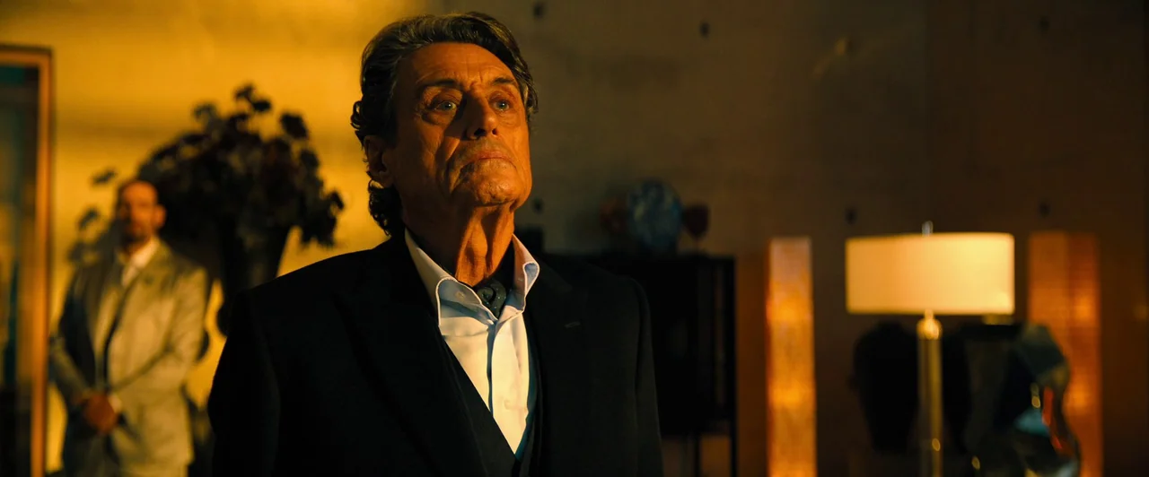John Wick: Chapter 4 (2023) movie still: medium — A spacious, dimly lit room contains modern furniture and warm glowing lamps. An older man in a…