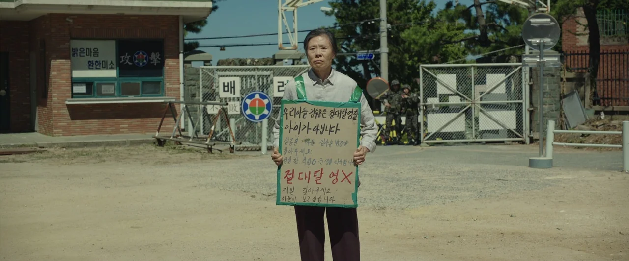 Along with the Gods: The Two Worlds (2017) movie still: medium — An elderly woman stands before a fenced military entrance with a brick guard…
