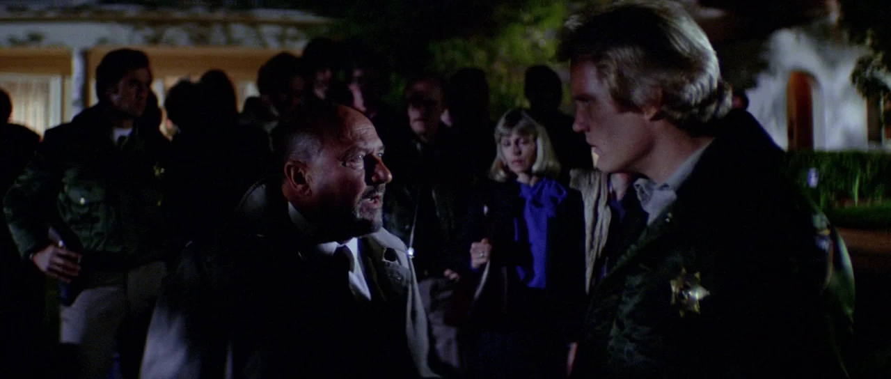 Halloween II (1981) movie still: medium — A dark residential street at night with a group of people gathered. A man and a uniformed officer talk…