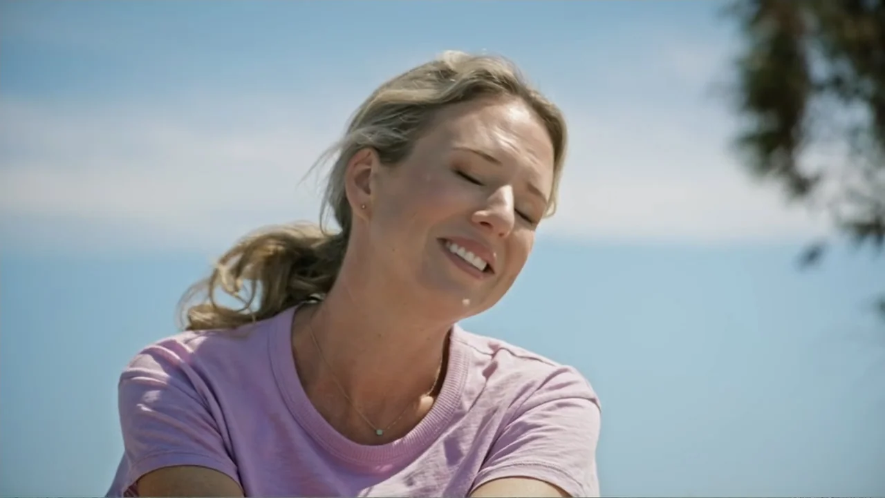 Making Waves (1998) movie still: medium — An outdoor setting features a clear bright blue sky in the background. A blonde woman in a pink t-shirt…