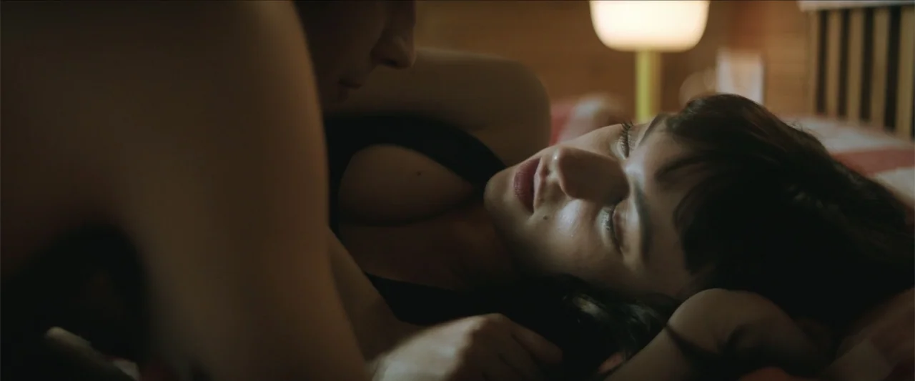 Tell Me Softly (2025) movie still: closeup — A dimly lit room features a person lying in bed next to another individual. A woman with dark hair…