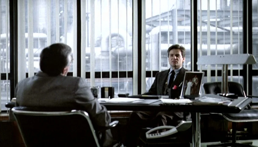 The Seventh Continent (1989) movie still: medium — An executive office features a large window with vertical blinds and an industrial backdrop. Two…