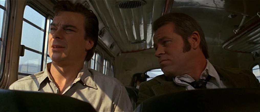 Assault on Precinct 13 (1976) movie still: medium — Interior of a moving bus with rounded windows and ribbed metallic ceiling details. Two men sit…