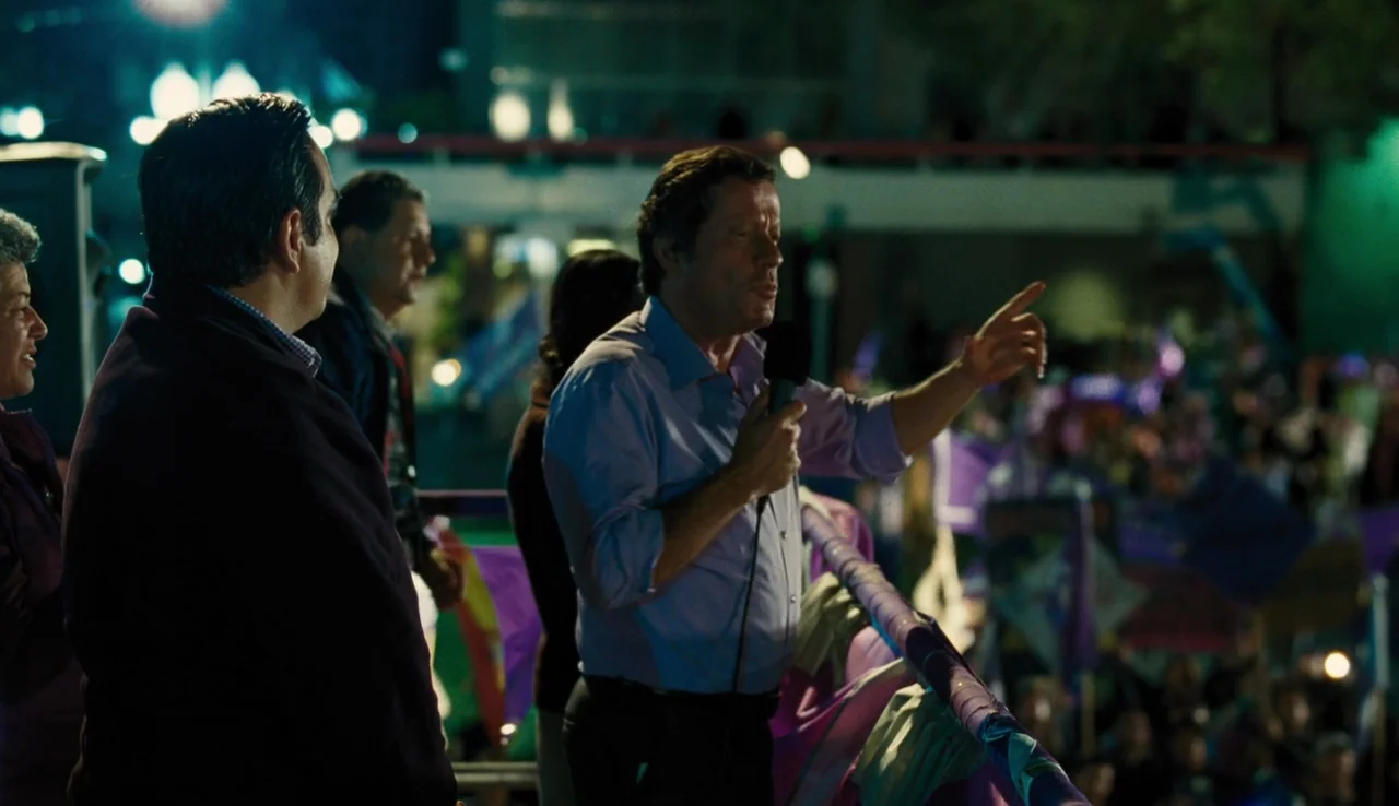 Our Brand is Crisis (2015) movie still: medium — A night rally platform with several people observing a speaker in the foreground. The speaker…