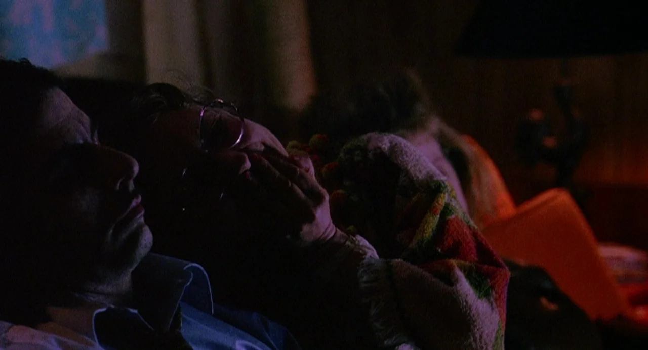 Office Killer (1997) movie still: medium — A dark, dimly lit room is illuminated by the glow of a television. Two men sit together while covered by…