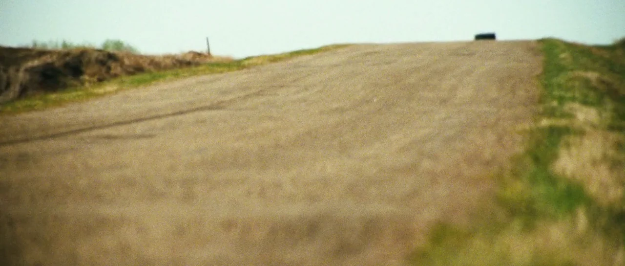 Surveillance (2008) movie still: establishing — A rural dirt road rises to meet the horizon under a bright sky. A dark vehicle is silhouetted at the…