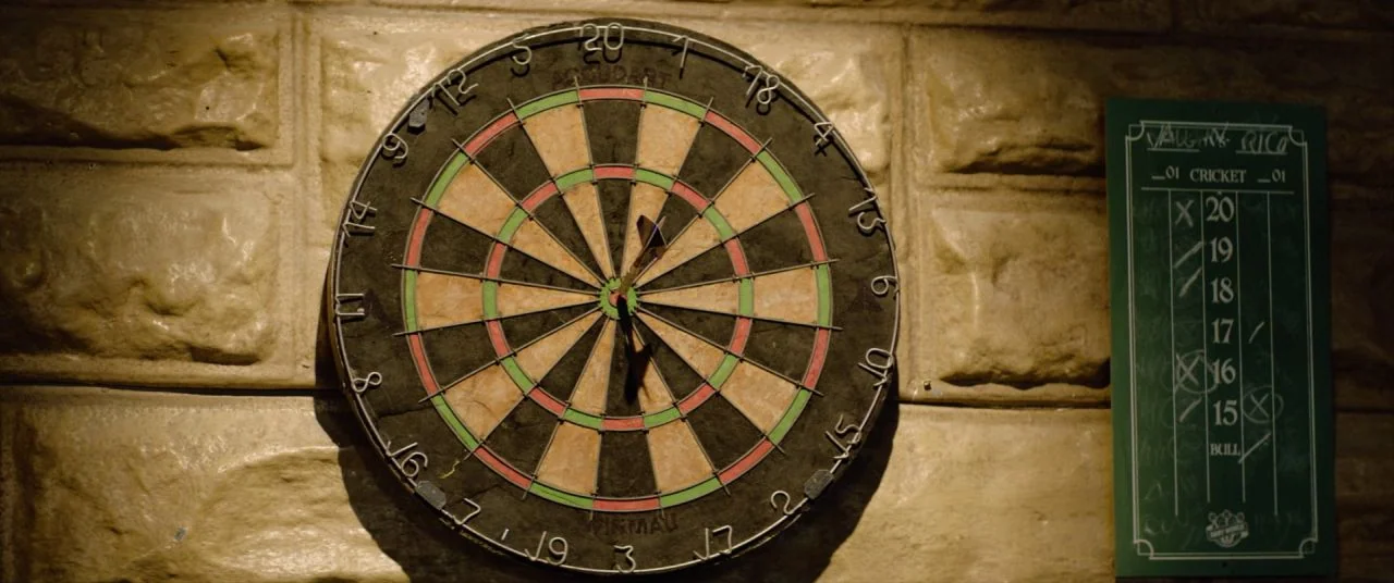 Palm Springs (2020) movie still: closeup — A wall composed of tan-colored stone blocks features a mounted dartboard and a scoreboard. A single dart…