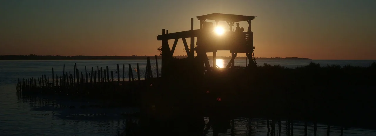 28 Years Later (2025) movie still: wide — A wooden fishing structure sits on stilts over calm water at sunset. The structure is rendered as a black…