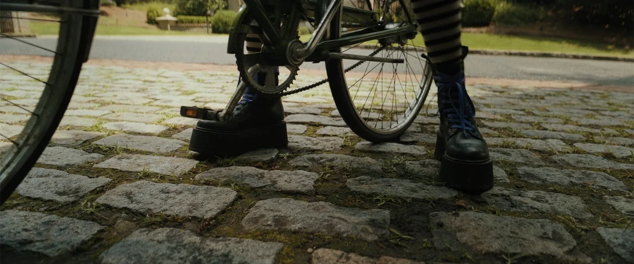 Black Adam (2022) movie still: medium — A close-up of a person's legs and feet standing on a bicycle on cobblestone. The person wears platform boots…