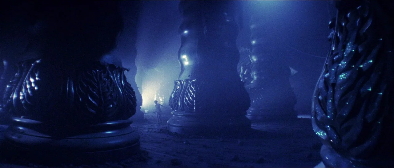 Legend (1985) movie still: wide — A dark subterranean chamber contains large, ornate stone pillars with intricate leaf-like carvings. The interior…