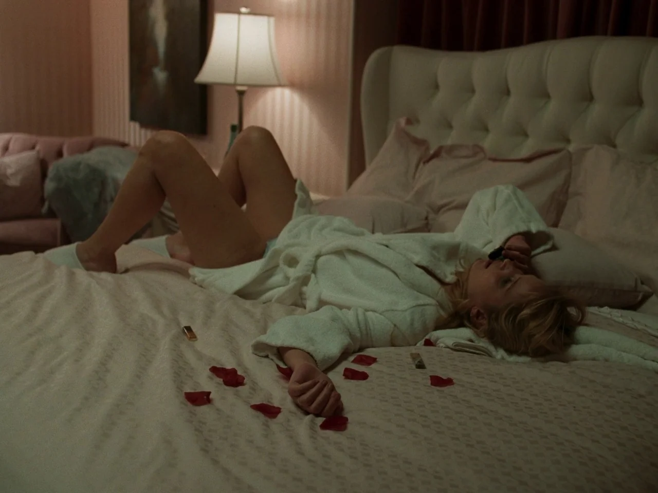 Die My Love (2025) movie still: medium — A well-appointed bedroom with striped wallpaper features a large tufted white headboard bed. A woman in a…