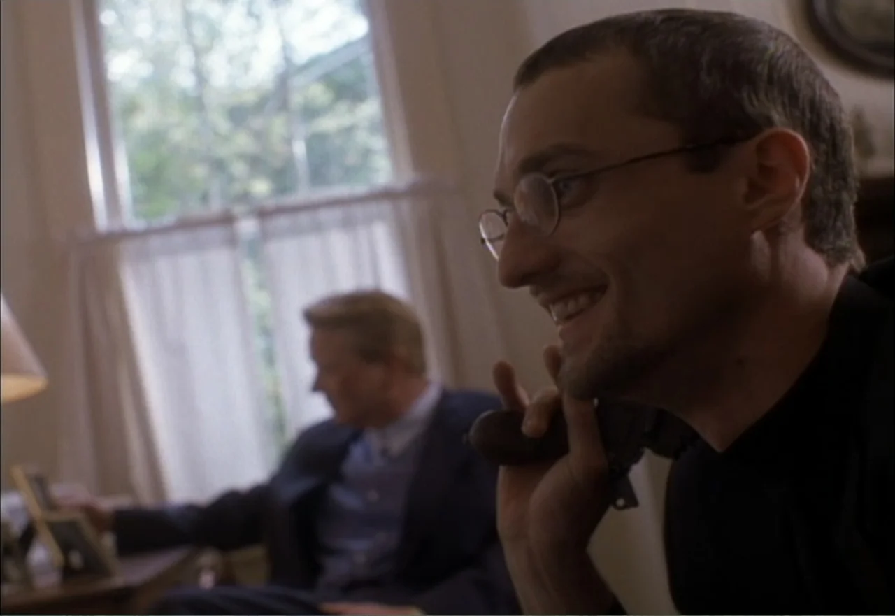 Ambushed (1998) movie still: closeup — A living room with a large window and white curtains in the background. A man holds a phone to his ear in the…
