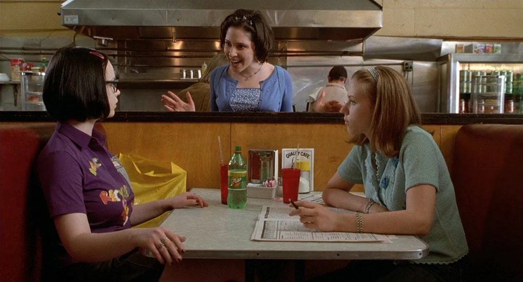 Ghost World (2001) movie still: medium — A diner booth with red vinyl seating and a wood-paneled wall backdrop. Three teenage girls sit together at…