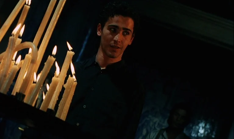 The Rose King (1986) movie still: medium — A dimly lit interior space features dark walls and a blurred figure in the background. A young man stands…