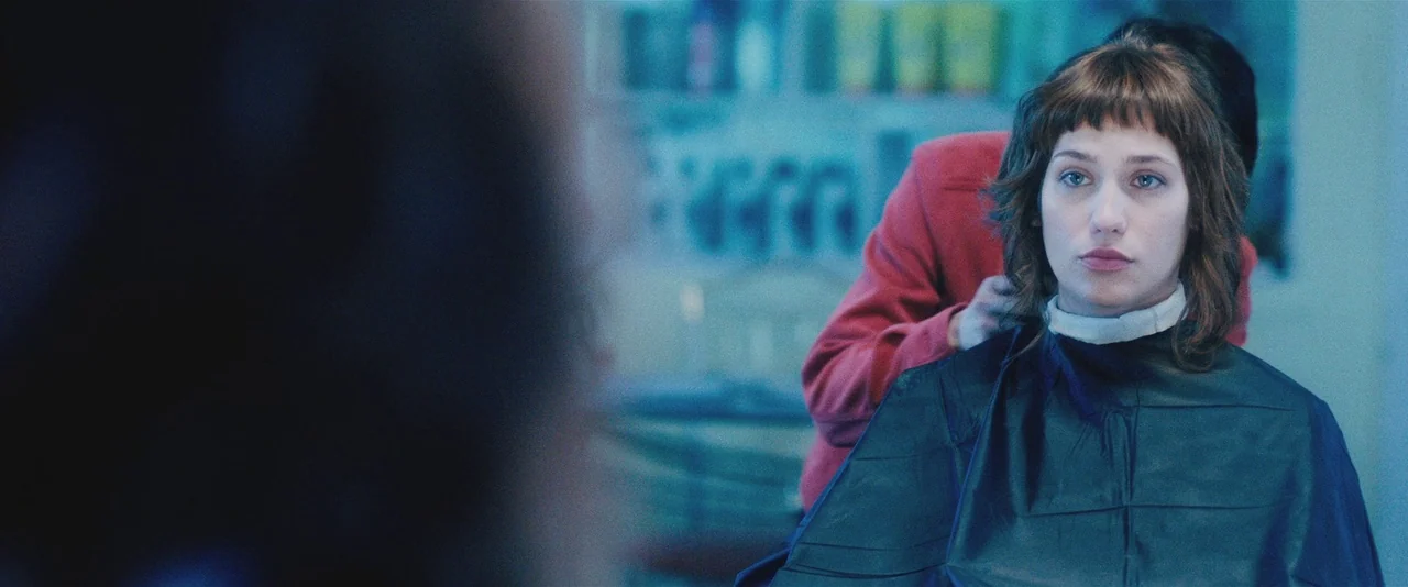 Gemini (2017) movie still: medium — A woman sits in a chair in a brightly lit professional hair salon interior. She wears a black protective cape…