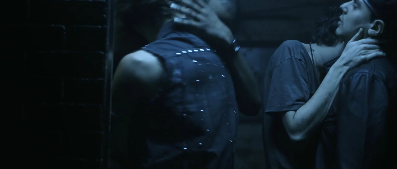 NY84 (2016) movie still: medium — A dark, dimly lit corner with a brick wall behind two young men. The two individuals embrace while standing close…