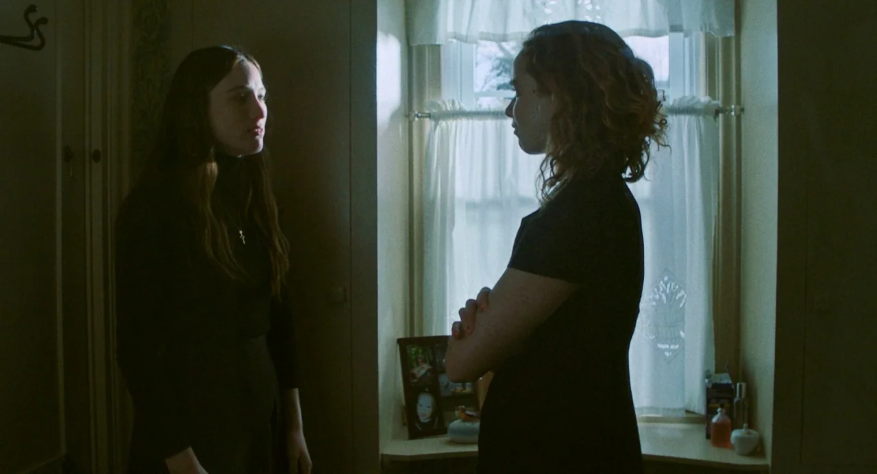 Blow The Man Down (2019) movie still: medium — A residential room features a window covered by white curtains and a side table. Two women stand in…