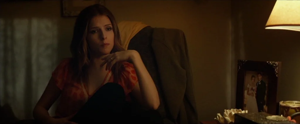 Cake (2014) movie still: medium — A dimly lit residential room features a woman sitting in a dark upholstered chair. To her right, a framed wedding…