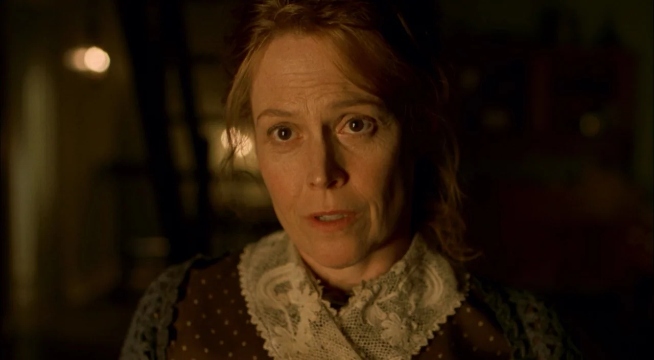The Village (2004) movie still: closeup — A dimly lit room with dark wooden furniture and soft, warm ambient light. A middle-aged woman with…