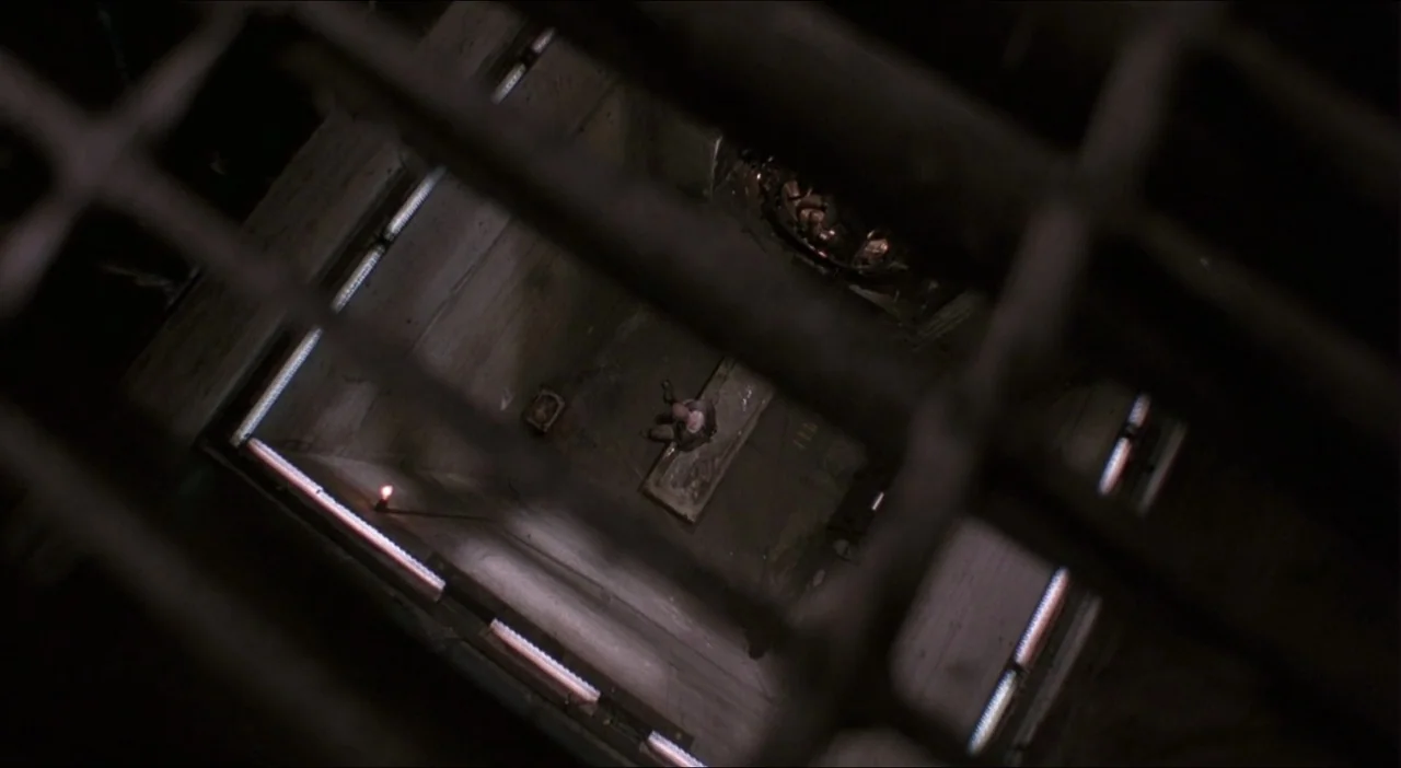 Twelve Monkeys (1995) movie still: wide — A stark, concrete-walled enclosure features a single raised cot and institutional lighting fixtures. A…