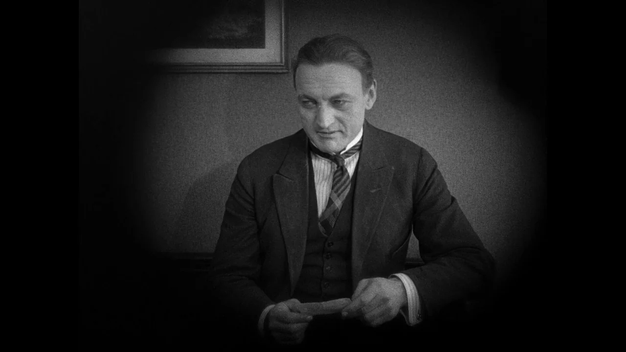 Master of the House (1925) movie still: medium — A man sits in a dimly lit room against a plain wall. He looks down at a small object held in his…