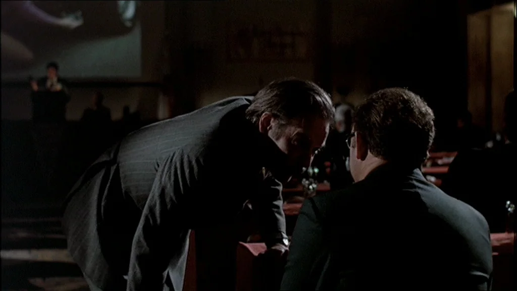 The Conversation (1974) movie still: medium — A dimly lit conference hall features rows of tables with red covers in darkness. A man in a pinstripe…