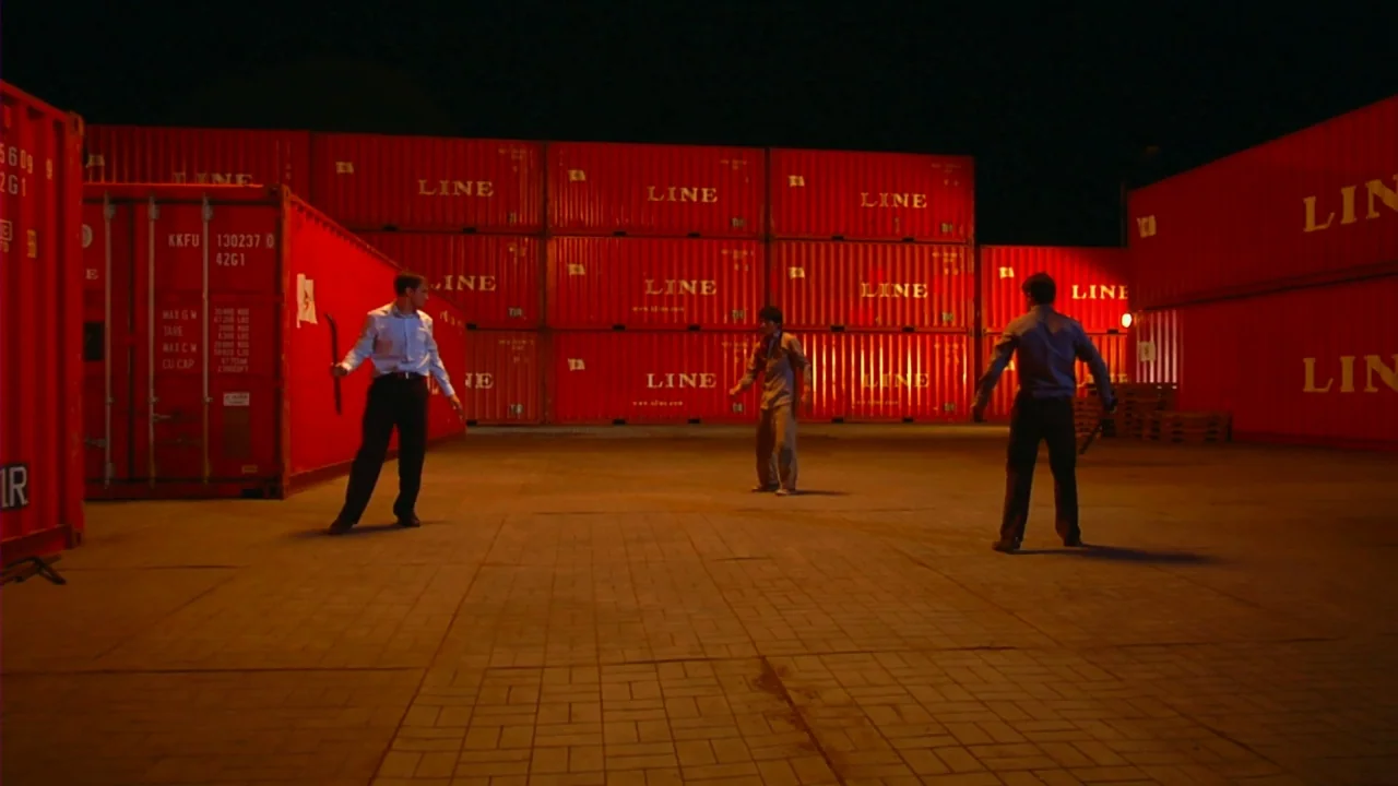 Merantau (2009) movie still: wide — A dark, paved yard surrounded by high stacks of bright red shipping containers at night. Three men stand apart…
