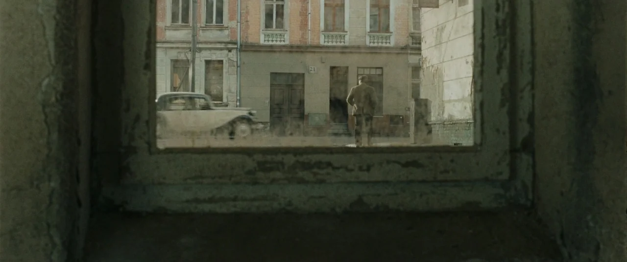 Phoenix (2014) movie still: wide — A view through a weathered, peeling window frame onto a paved urban street. A man in a wool coat stands alone on…