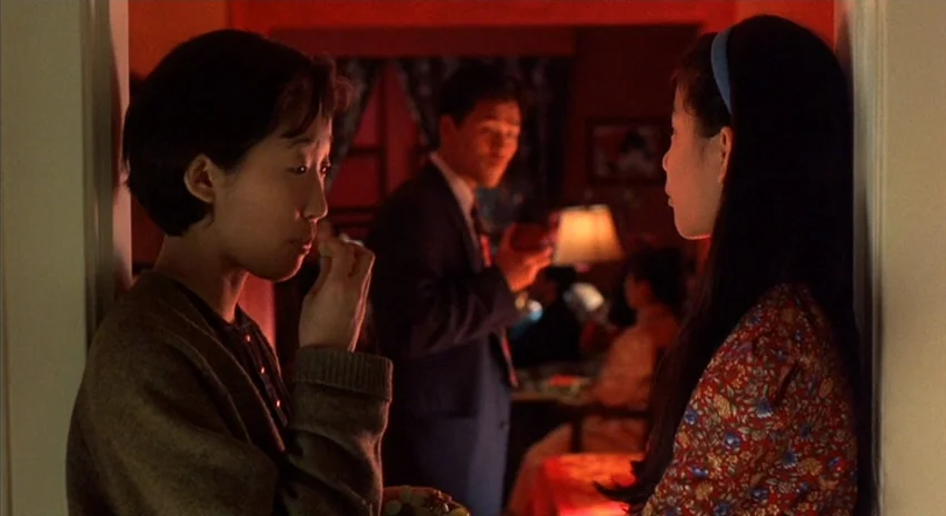 Double Happiness (1994) movie still: medium — A dimly lit room with deep red walls and a vintage table lamp. Two women in the foreground share…