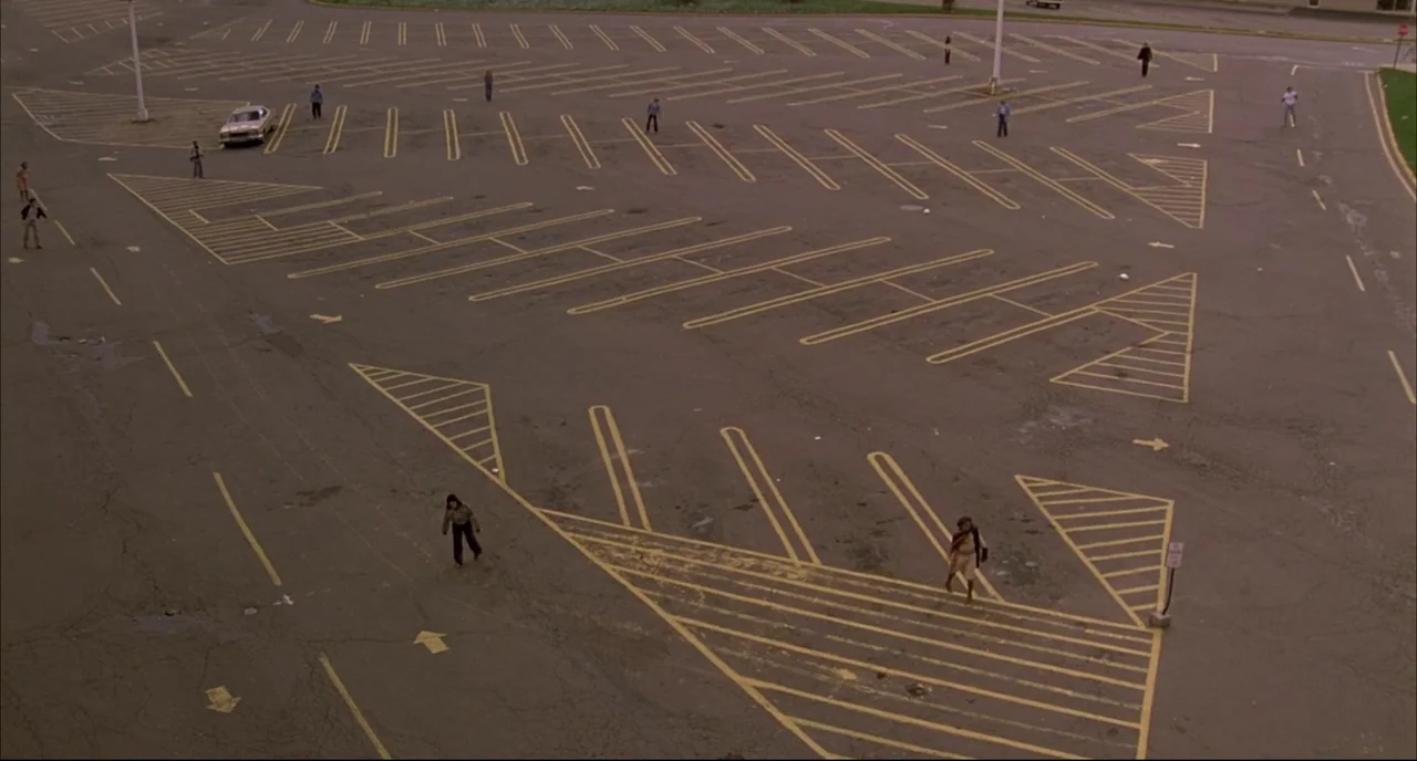 Dawn of the Dead (1978) movie still: wide — A vast, cracked asphalt parking lot is marked with faded yellow traffic paint lines. Multiple scattered…