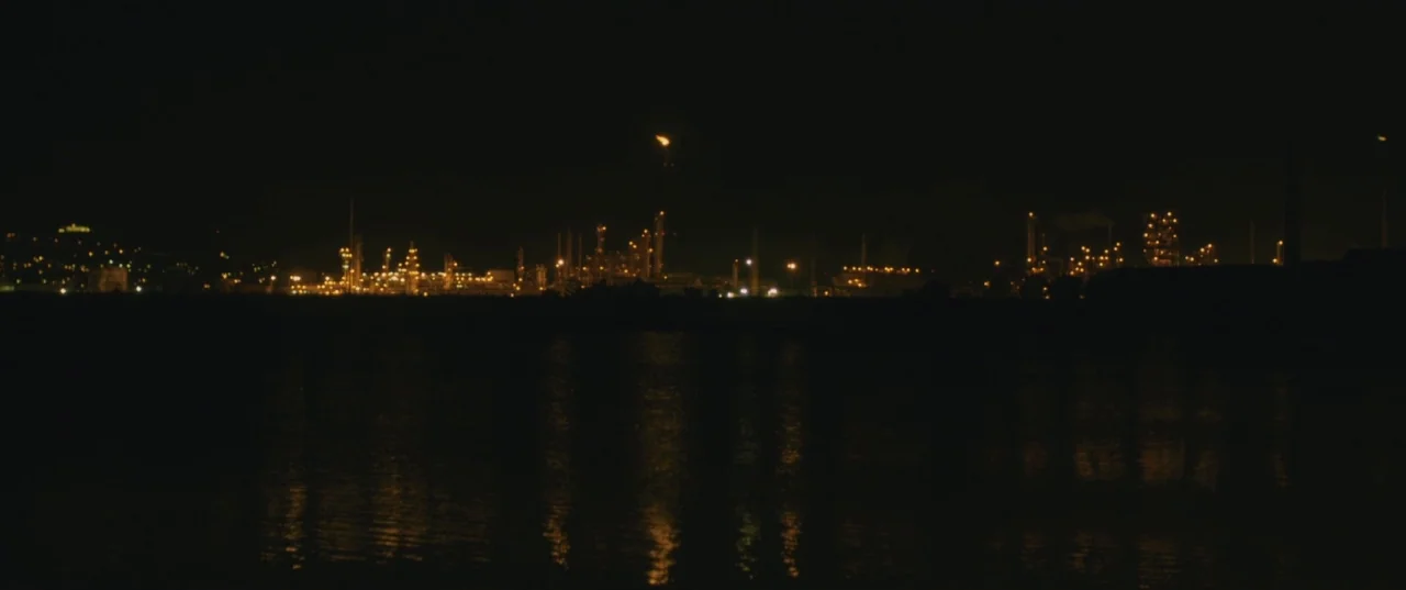 Evolution (2015) movie still: establishing — An industrial facility glows at night across a dark, calm body of water. The sprawling complex features…