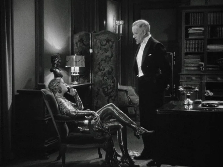 Mata Hari (1931) movie still: medium — A wood-paneled room contains a desk, a bookshelf, and a decorative privacy screen. A man stands formally near…