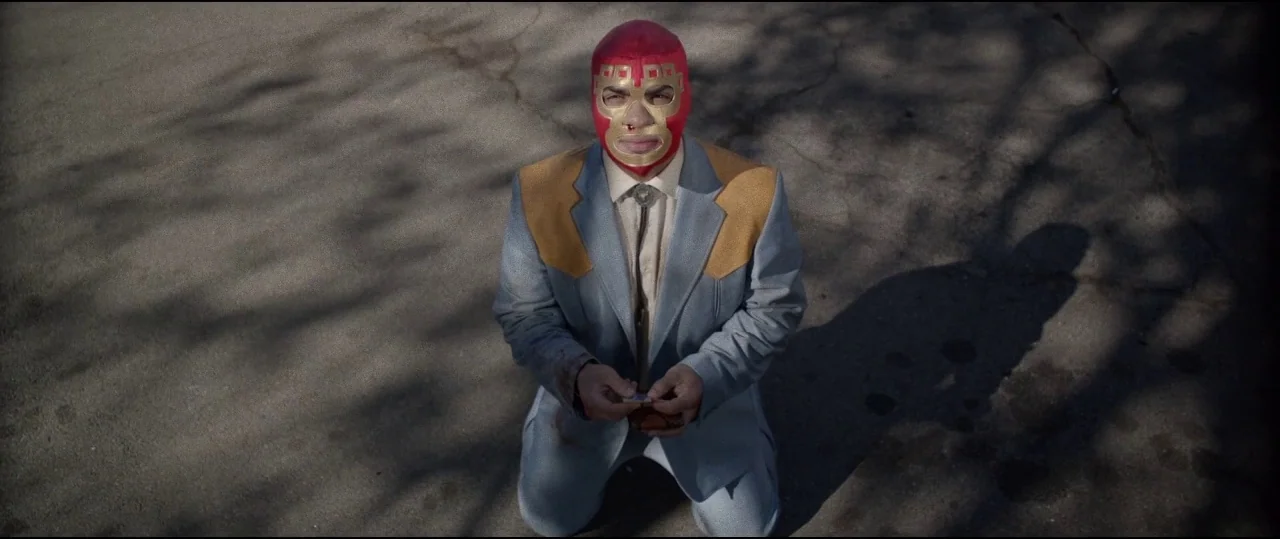 Lowlife (2017) movie still: medium — An expanse of gray asphalt marked by dark, long shadows from unseen trees. A man wearing a red wrestling mask…