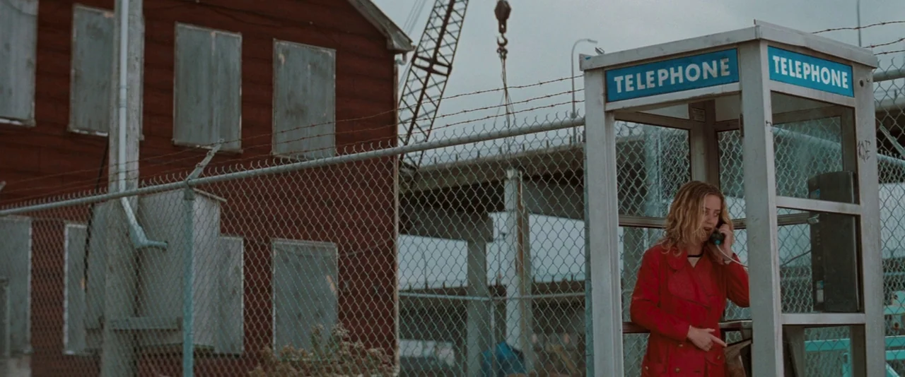 The Sweet Hereafter (1997) movie still: medium — A desolate industrial area features a fenced-off red building with boarded windows. A woman wearing…