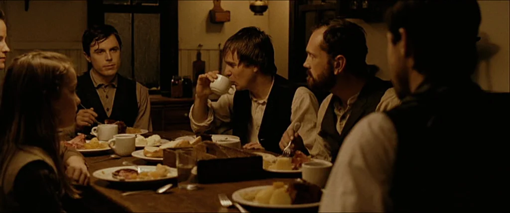 The Assassination Of Jesse James By The Coward Robert Ford (2007) movie still: medium — Several people sit around a long wooden table in a dimly…