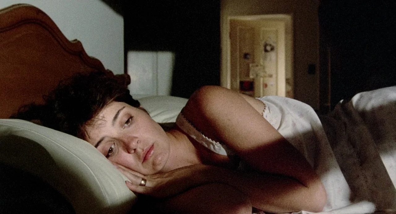 Crimes of Passion (1984) movie still: closeup — A dimly lit room features a woman lying on a bed. She rests her head on her hand against a white…