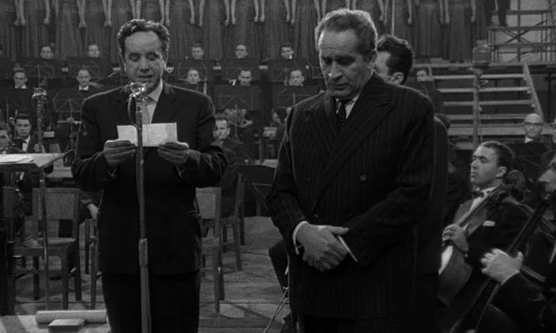 Man Is Not Bird (1965) movie still: medium — A dimly lit orchestral stage featuring rows of seated musicians in the background. Two men in formal…