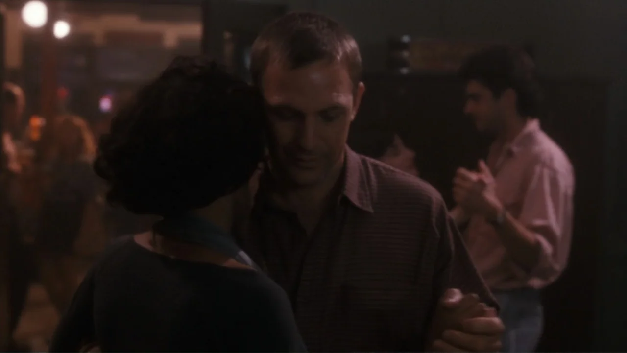 The Bodyguard (1992) movie still: medium — A dimly lit indoor bar setting with blurred figures in the background. A man and a woman stand face to…
