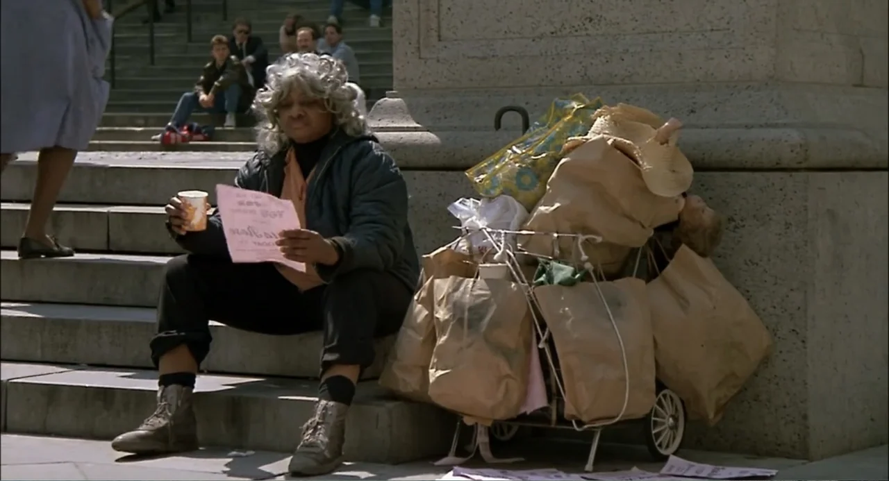 She-Devil (1989) movie still: medium — A woman sits on large stone steps next to a cart piled with bags. She holds a paper pamphlet and a beverage…