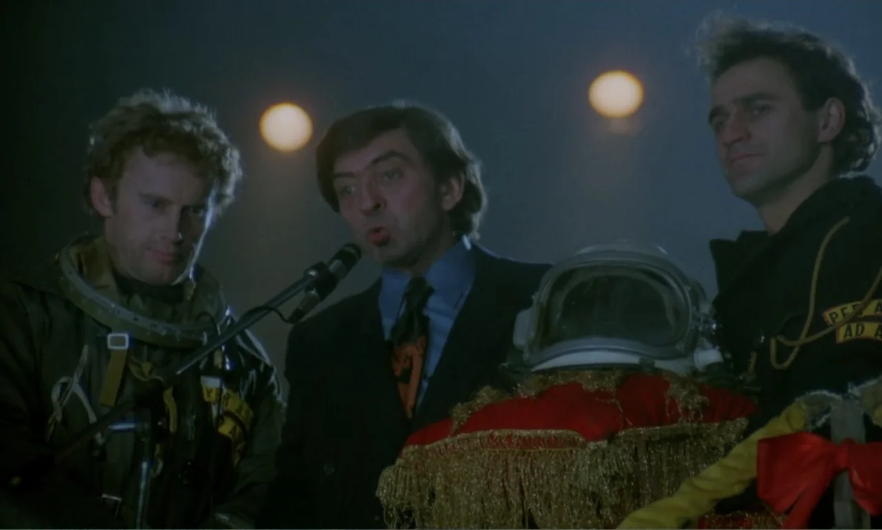 Ga-ga: Glory to the Heroes (1986) movie still: medium — Three men stand outdoors at night behind a microphone near a large space helmet. A man in a…